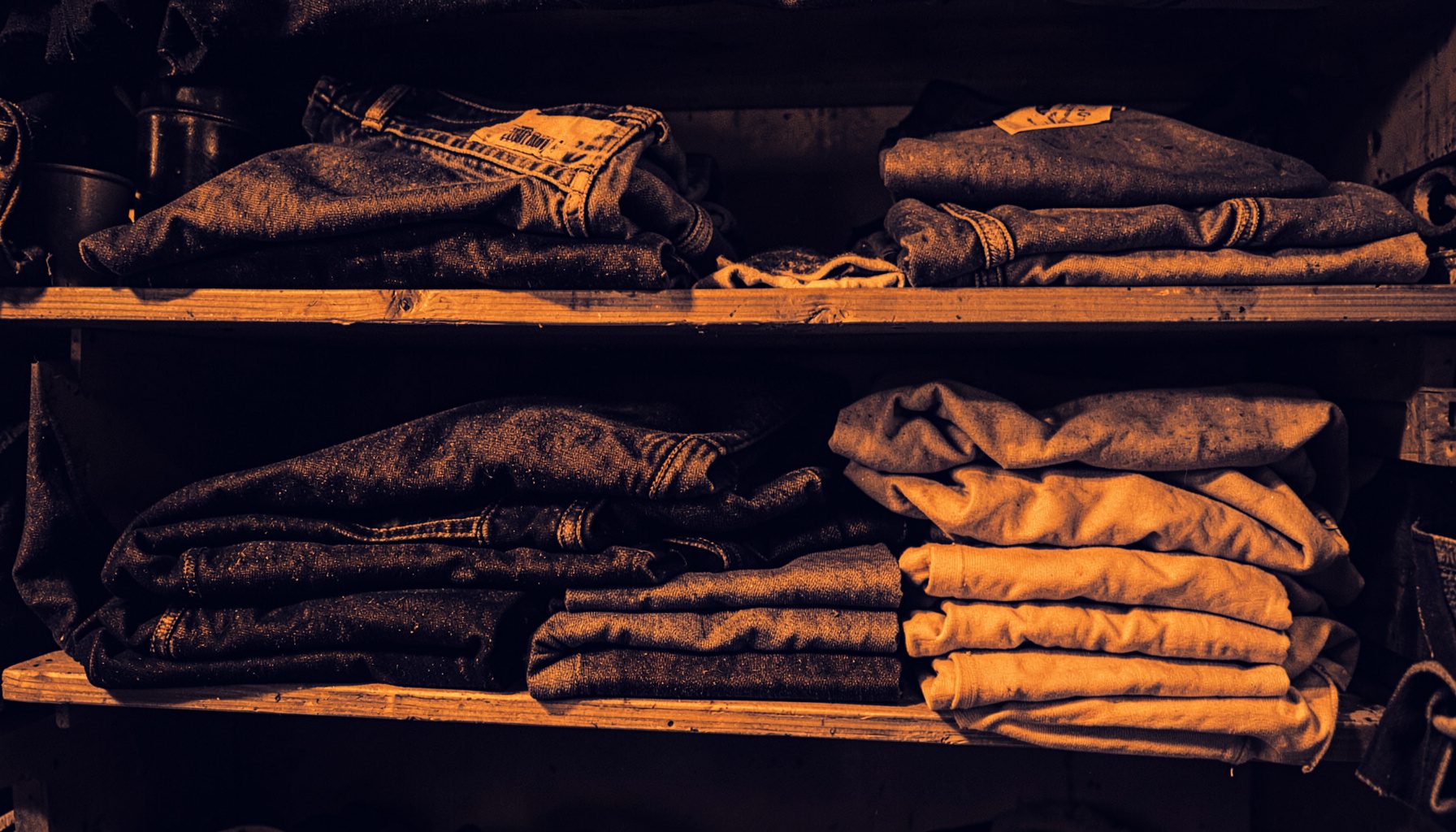 A duotone photograph in deep navy and warm amber tones showing folded denim jeans and cotton t-shirts stacked neatly on wooden shelves in a workshop.