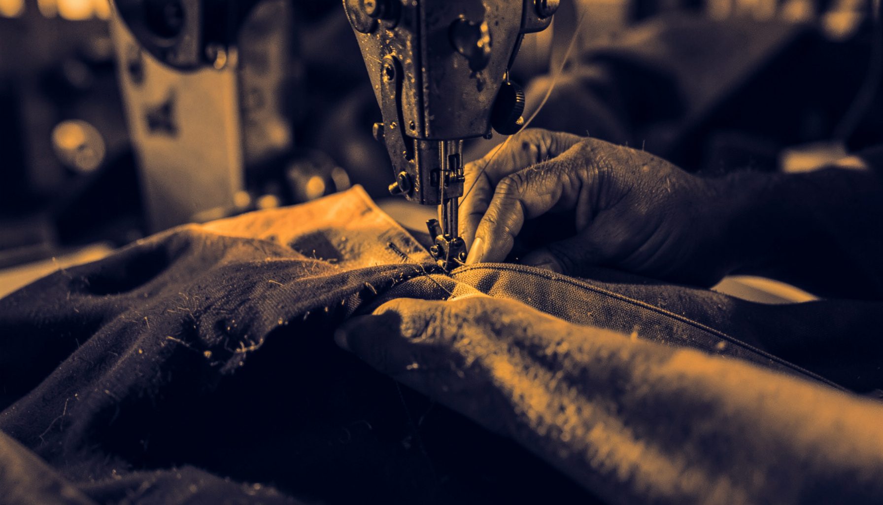 A duotone photograph in deep navy and warm amber tones showing a craftsman's hands stitching heavy canvas fabric on an industrial sewing machine.