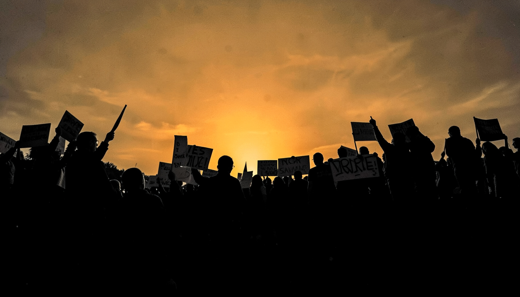 A wide atmospheric photograph of a labor rally at dusk, rendered as a hard two-tone duotone in deep charcoal black and warm amber gold, with silhouetted figures holding signs against a glowing sky.